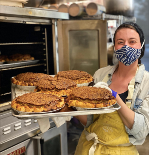 A smiling person in a mask holds a tray of freshly baked pies in a kitchen, with an oven in the background.
