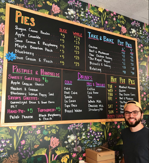A man stands next to a colorful menu board featuring pies, pastries, drinks, and specials against a floral background.