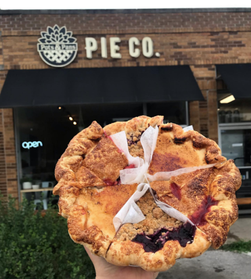 A person holds a large, messy pie outside a pie shop with a sign that reads "Pie Co." in the background.