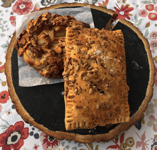 Two baked pastries on a wooden platter: one with a flaky crust and seeds, the other topped with almonds and fruit.