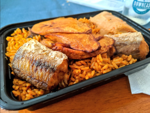 A takeout container filled with rice, fried plantains, and grilled fish, set on a wooden table.