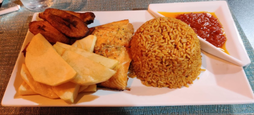 A plate of jollof rice, grilled fish, fried plantains, and sliced apples, served with a side of spicy sauce.