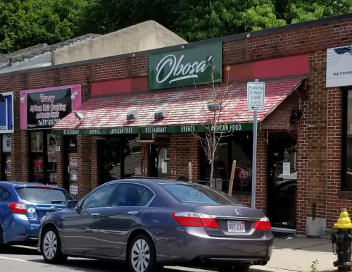 A brick restaurant facade with a sign reading "Obosa" and parked cars in front, featuring a tree and parking sign.