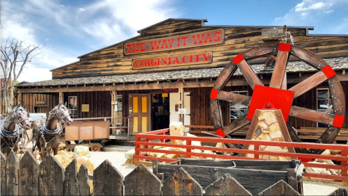A rustic wooden building with a large red wheel outside, featuring a sign that reads "The Way It Was, Virginia City."