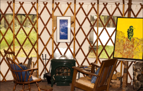 Cozy interior of a yurt with wooden furniture, a green stove, and artwork against a backdrop of large windows.