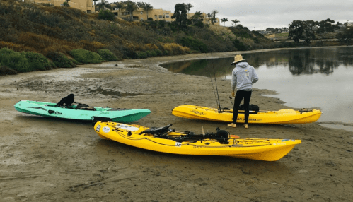 A person in a hat stands near two kayaks on a sandy shore, with a calm water backdrop and greenery in the distance.