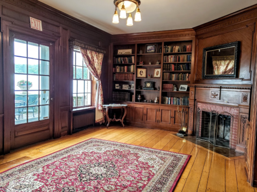 Cozy room with wooden walls, a bookshelf, a fireplace, and a large window overlooking a scenic view.