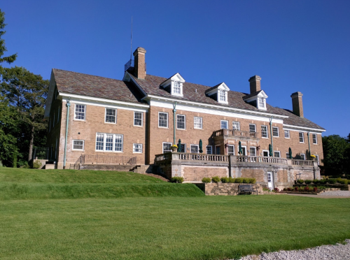 A large brick mansion with multiple chimneys, surrounded by green grass and trees under a clear blue sky.