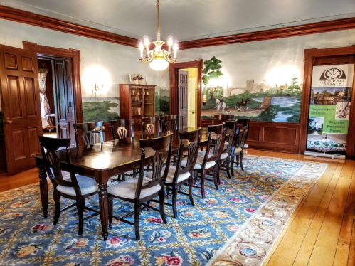 A spacious dining room with a long wooden table, chairs, and a decorative rug, featuring a mural and a banner in the corner.