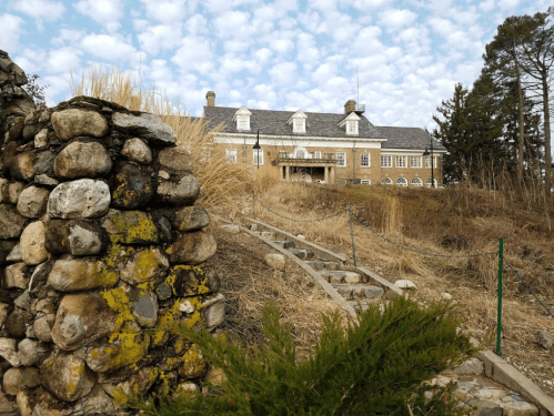 A stone pathway leads to a large, historic building surrounded by tall grass and a cloudy sky.