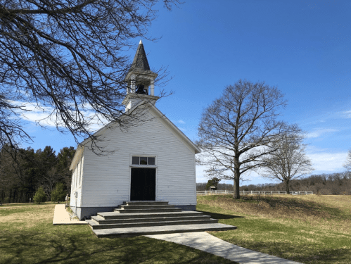 A white church with a steeple, surrounded by grass and trees, under a clear blue sky.