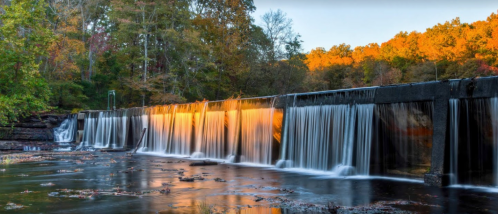 A serene waterfall cascades over a dam, surrounded by colorful autumn foliage and a calm river.