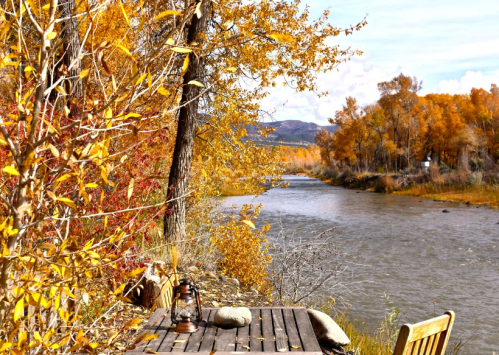 A serene riverside scene with autumn foliage, a wooden table, and a lantern beside a gently flowing river.