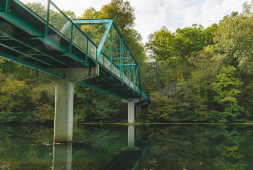 A green metal bridge spans over a calm, reflective body of water, surrounded by lush trees and a cloudy sky.