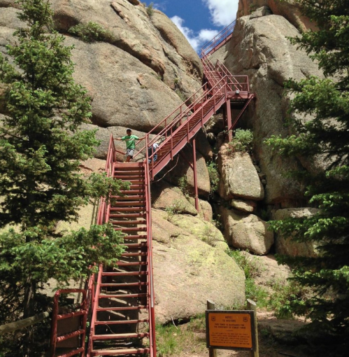 A steep metal staircase leads up between large rock formations, surrounded by trees and a sign at the base.