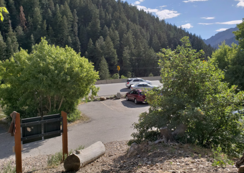 A view of a parking area surrounded by trees, with a road and mountains in the background under a clear blue sky.
