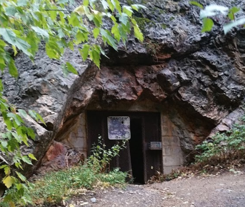 A gated entrance to a cave or mine, surrounded by rocky terrain and greenery. A warning sign is visible on the gate.
