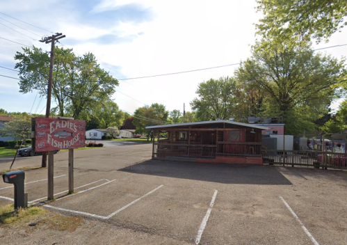 A rustic fish house with a sign, surrounded by trees and an empty parking lot on a sunny day.