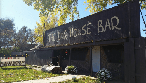 Exterior of The Dog House Bar with a wooden sign, open Tuesday to Saturday, surrounded by trees and flowers.