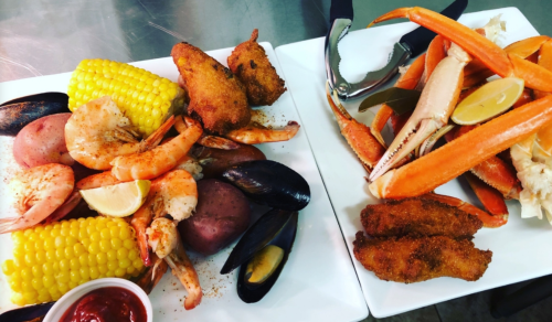 A seafood platter featuring shrimp, crab, mussels, corn, potatoes, and fried fish, served with lemon and dipping sauce.
