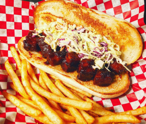 A toasted sandwich with BBQ meat and coleslaw, served with a side of golden French fries in a red-checkered basket.