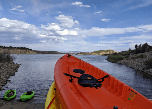 An orange kayak on a shore with two green kayaks nearby, set against a scenic lake and cloudy sky.