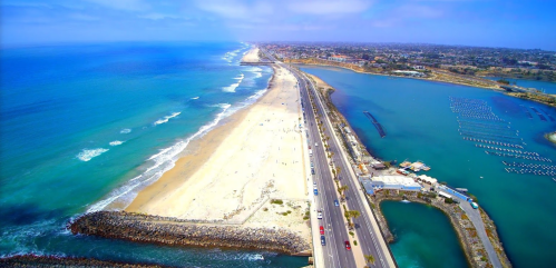Aerial view of a coastal road along a sandy beach, with blue ocean on one side and a marina on the other.