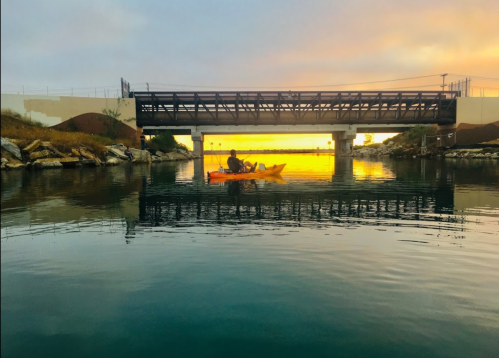 A person kayaking under a bridge at sunset, with calm water reflecting the colorful sky.