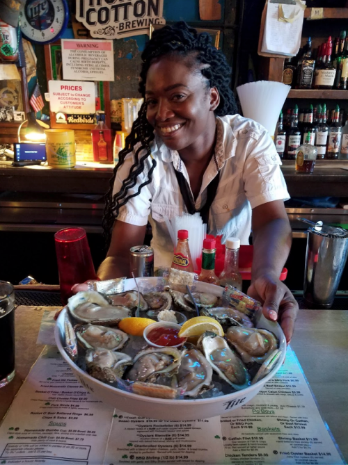 A smiling woman serves a large platter of oysters at a bar, surrounded by drinks and condiments.