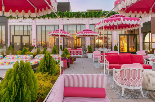 A vibrant rooftop terrace with pink umbrellas, colorful seating, and lush greenery, creating a cheerful atmosphere.