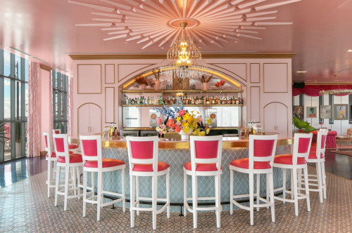 A stylish bar with pink decor, white stools, and a vibrant floral arrangement, featuring a chandelier and shelves of drinks.