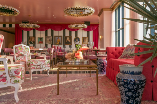 A stylish lounge with floral chairs, a glass table, and vibrant decor, featuring artwork on the walls and large windows.