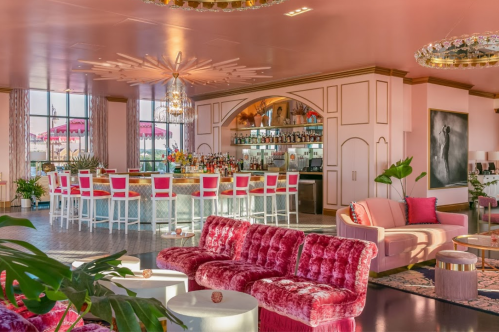 A stylish bar with pink decor, plush seating, and vibrant flowers, featuring a well-stocked bar and elegant lighting.