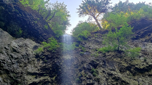 A waterfall cascades down rocky cliffs, surrounded by lush green trees and a clear blue sky above.