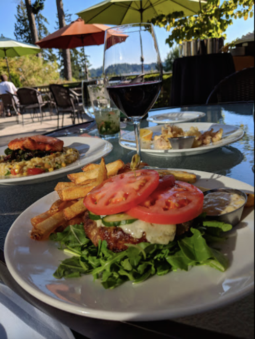 A plate with a burger topped with tomatoes and greens, fries, and a glass of red wine, set outdoors with umbrellas.