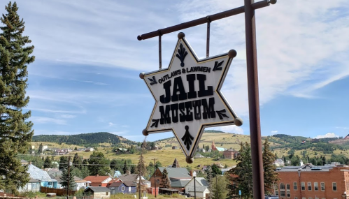 Sign for the Outlaws & Lawmen Jail Museum, featuring a star shape, against a backdrop of hills and buildings.