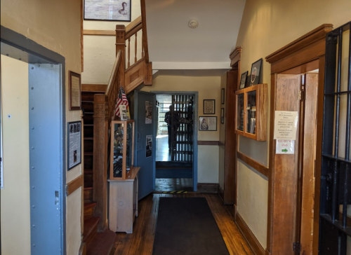 A hallway with wooden floors, framed photos on the walls, and a staircase leading to an upper level. A barred door is visible.