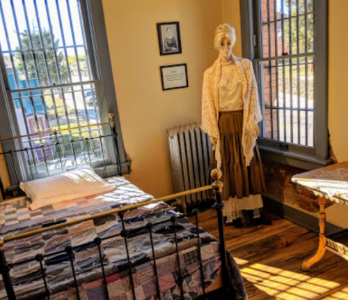 A vintage bedroom with a bed, a mannequin in period clothing, and large windows with bars.