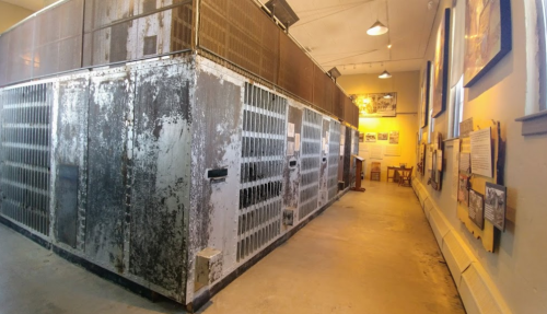 Interior view of a historic prison cell block with metal bars, peeling paint, and informational displays on the walls.