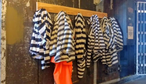 A row of black and white striped prison uniforms hanging on a wooden rack against a weathered wall.