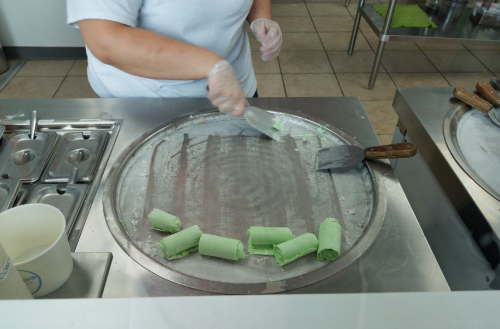 A person prepares rolled ice cream on a cold metal surface, with green ice cream being shaped into rolls.