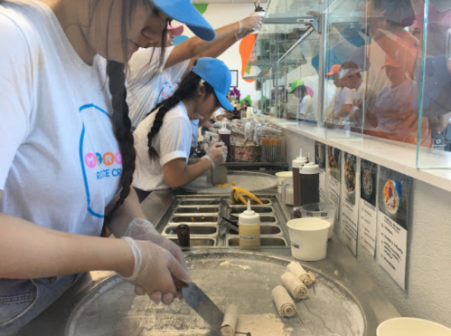 Two employees make rolled ice cream at a counter, with various toppings and tools visible in the background.