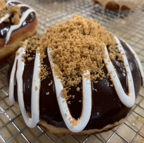 A chocolate frosted donut topped with crumbled cookie and white icing drizzles, resting on a cooling rack.