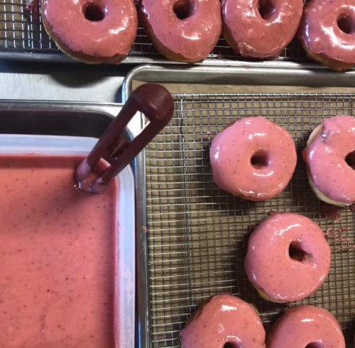 Freshly glazed pink donuts on a cooling rack, with a bowl of pink icing nearby.