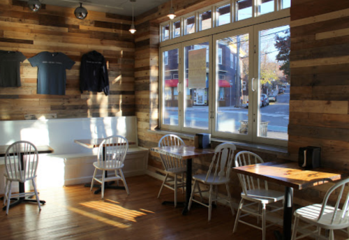 Cozy café interior with wooden walls, white chairs, and tables, featuring sunlight streaming through large windows.