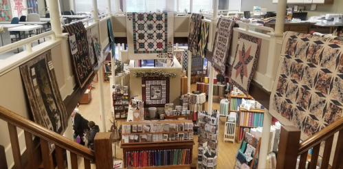 A spacious fabric store with colorful quilts hanging, shelves of fabric, and customers browsing below.