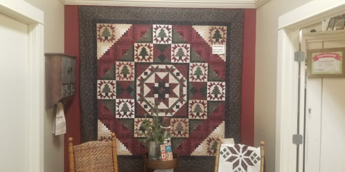 A colorful quilt hangs on a wall, surrounded by two chairs and a small table with decorative items.