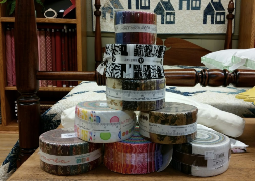 A stack of colorful fabric rolls and jelly rolls arranged on a table, with a quilted backdrop.