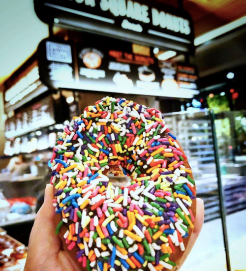 A hand holding a colorful donut covered in rainbow sprinkles, with a bakery sign in the background.