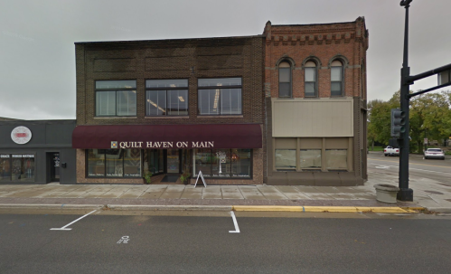 A two-story building with a maroon awning labeled "Quilt Haven on Main" and large windows, located on a street corner.